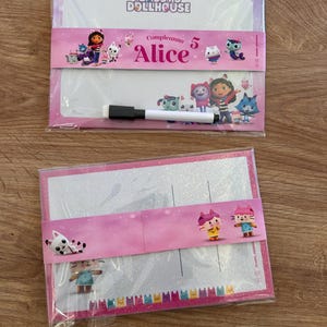 Puede incluir: Dos pizarras blancas rectangulares con la marca Gabby's Dollhouse. La pizarra superior incluye un marcador negro y una banda de papel rosa con el texto 'Compleanno 5 Alice' y personajes. La inferior tiene una banda rosa con personajes gatunos.