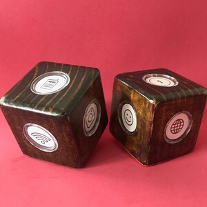 Può includere: Due dadi di legno con una macchia scura e inserti circolari trasparenti. Ogni dado presenta diverse icone bianche, tra cui un menu, una faccina sorridente, un simbolo wifi, un globo e un bersaglio. I dadi sono su uno sfondo rosso pieno.