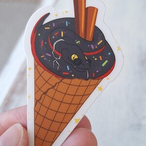May include: A sticker depicting a cartoon snake in an ice cream cone. The snake is black with yellow eyes and a red accent. The ice cream is black with colourful sprinkles, and the cone is brown with a waffle pattern. Two brown sticks are in the ice cream.