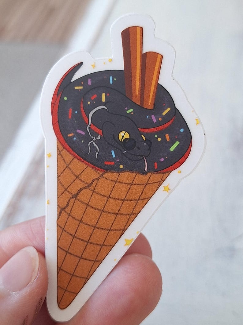 May include: A sticker depicting a cartoon snake in an ice cream cone. The snake is black with yellow eyes and a red accent. The ice cream is black with colourful sprinkles, and the cone is brown with a waffle pattern. Two brown sticks are in the ice cream.