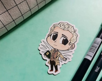 May include: A sticker of a cartoon character with blonde hair, large brown eyes, and angel wings. The character wears a tan coat and holds a green book. The sticker is on a light green surface with black pens.