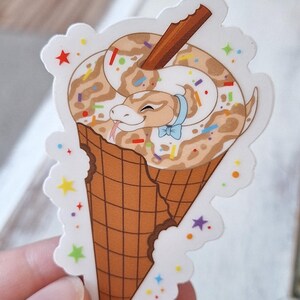 May include: A sticker depicting a cartoon snake in a chocolate ice cream cone. The snake has a light brown and white pattern, a blue bow tie, and a cinnamon stick. The cone is brown with a bite taken out, and the sticker has a white border with colourful stars.