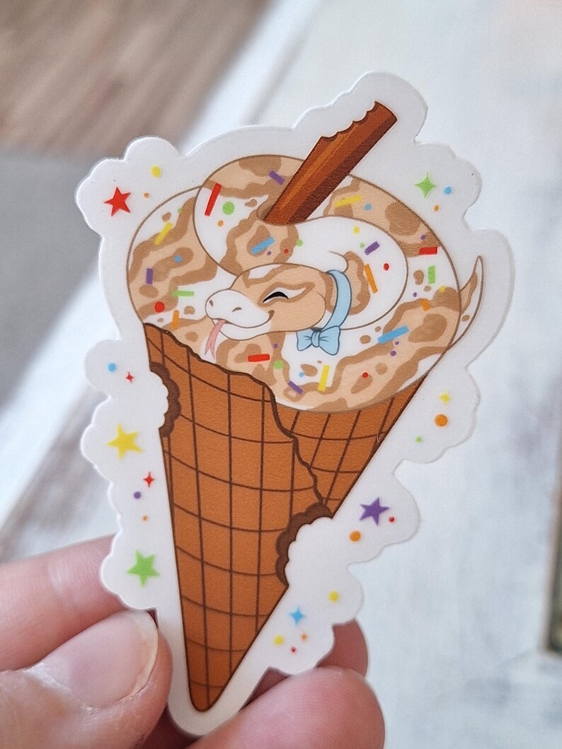 May include: A sticker depicting a cartoon snake in a chocolate ice cream cone. The snake has a light brown and white pattern, a blue bow tie, and a cinnamon stick. The cone is brown with a bite taken out, and the sticker has a white border with colourful stars.