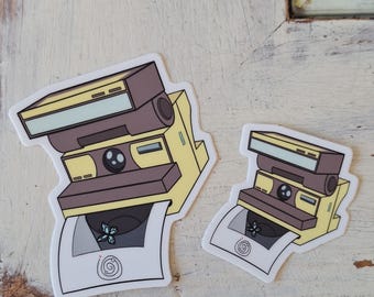 May include: Two stickers of a vintage Polaroid camera in yellow and brown. The camera is depicted with a photo being ejected, featuring a floral design and a spiral. The stickers have a white border.