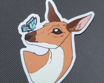 May include: A sticker depicting a deer's head with a butterfly. The deer is brown with white and pink details, and the butterfly is blue. The sticker has a white border and is on a textured grey surface.