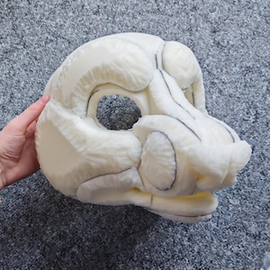 May include: A cream-coloured, unfinished fursuit head base, featuring a stylised animal design. The head has large eye openings, a prominent snout, and detailed facial features outlined in black. The base is made of a soft, textured material.