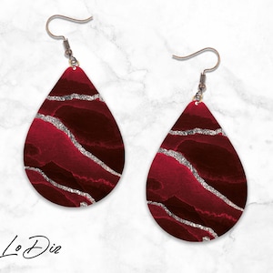 May include: A pair of teardrop-shaped earrings with a red and silver abstract design. The earrings have silver glitter accents and are suspended from silver hooks.