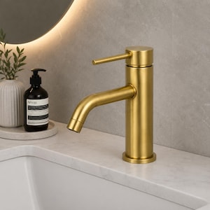 Puede incluir: Un grifo de baño en oro cepillado con un mango cilíndrico único y un caño curvo. El grifo presenta un acabado metálico mate y un diseño sencillo, colocado sobre una encimera blanca junto a una botella de jabón de color marrón oscuro.