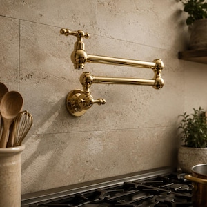 May include: A wall-mounted pot filler faucet with a polished gold finish, featuring a double-jointed articulating arm and a cross-handle design. The fixture is mounted on a light-colored stone tile backsplash above a black stovetop surface.