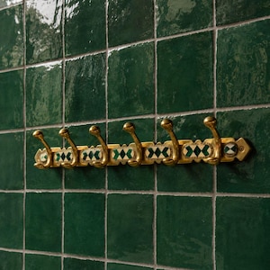 May include: A horizontal brass wall rack featuring six curved hooks. The central bar is decorated with repeating rectangular inlays of white and dark green geometric diamond patterns, secured to the wall with visible screws at both ends.