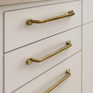 May include: Three horizontal brass drawer pulls with a polished finish and rounded ends, each secured to a flat-fronted white cabinet drawer by two visible screws at the mounting points, creating a clean and functional hardware arrangement.