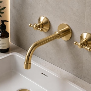 Könnte beinhalten: Eine wandmontierte Badezimmerarmatur mit gebürstetem Gold-Finish, bestehend aus einem geschwungenen Auslauf und zwei separaten Kreuzgriff-Bedienelementen. Die Armatur ist über einem weißen Keramikwaschbecken mit runden Rosetten installiert.