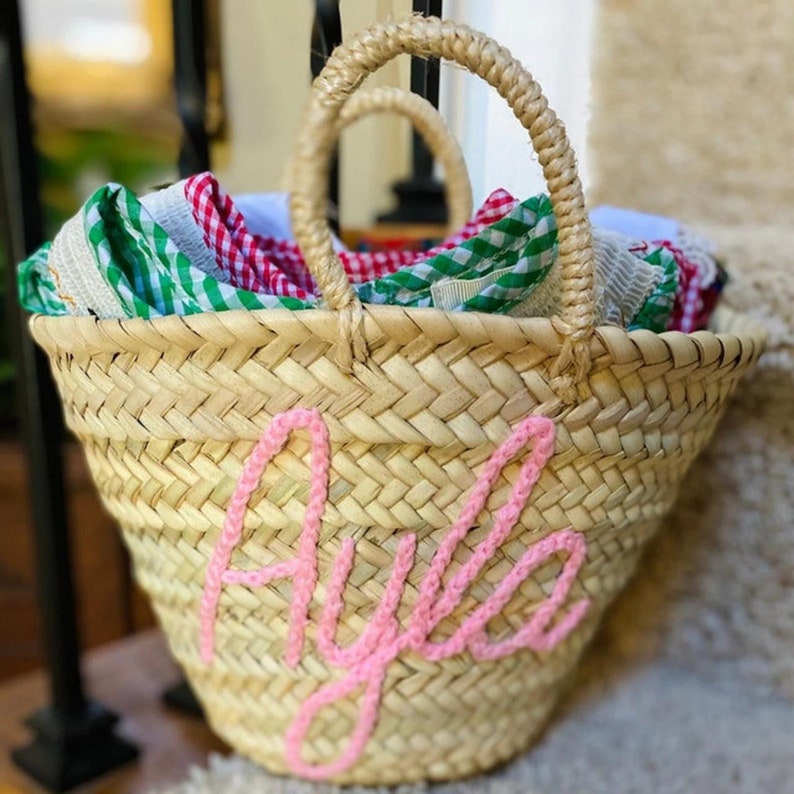 Op de afbeelding: Een strooien tas met de naam "Ayla" in roze gehaakt op de voorkant. De tas is gevuld met kleurrijk gingham-stof.