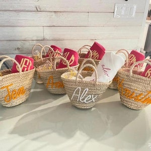 May include: A collection of woven straw tote bags with handles, each embroidered with a name in a contrasting color. Some bags contain folded pink towels or white shirts. The bags are arranged on a light-colored surface.