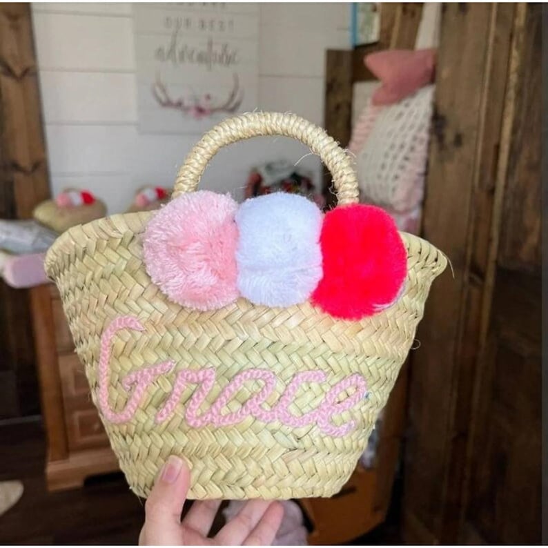 Op de afbeelding: Een kleine strooien tas met een roze, witte en rode pompon rand. De tas heeft een roze geborduurde naam "Grace" aan de voorkant.