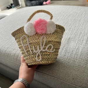May include: A small, woven straw basket with a rounded shape and a woven handle. The basket is decorated with three fluffy pom-poms in white and pink. The name "Ayla" is embroidered in white cursive on the front.