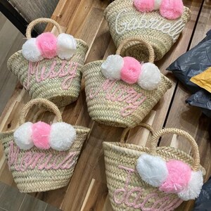 May include: Several woven straw tote bags with rounded handles are displayed. Each bag features a name embroidered in pink thread, adorned with pink and white pom-poms. The bags are arranged on a wooden surface.