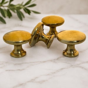 May include: A set of four round cabinet knobs made of polished brass with a smooth, reflective surface. Each knob features a flat circular top and a tapered base that connects to a circular mounting plate, showcasing a consistent metallic gold finish.