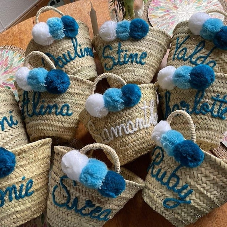 May include: Woven straw tote bags with handles, each personalized with a name in blue or white script. Each bag is adorned with a cluster of three pom-poms in shades of white, light blue, and dark blue. The bags are arranged on a wooden surface.