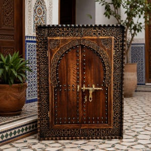 Puede incluir: Un armario rectangular de madera con dos puertas de diseño arqueado en la parte superior. La madera marrón oscura presenta tallas geométricas intrincadas en el marco e incluye un pestillo de latón y pequeños remaches metálicos en los bordes.