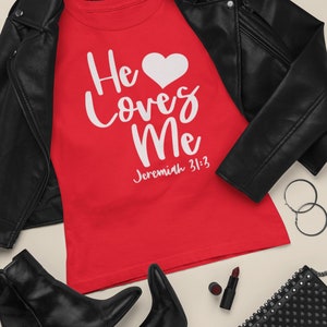 Puede incluir: Camiseta roja con texto blanco que dice "He loves Me" con un símbolo de corazón y la escritura "Jeremías 31:3". La camiseta está colocada sobre una superficie marrón claro con una chaqueta de cuero negra, botas negras, un bolso negro con tachuelas plateadas y un pintalabios rojo.