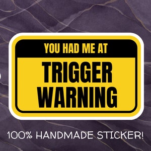 May include: Yellow and black warning sign sticker with the text "You had me at trigger warning" and the text "100% handmade sticker!" below the sign.