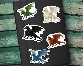 May include: A collection of fantasy-themed stickers featuring various dragons. The stickers showcase dragons in green, white, black, red, and blue, each with detailed wings and scales. The stickers are placed on a black surface.