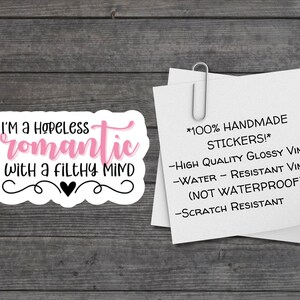 May include: A white sticker with a pink and black design. The sticker says "I'm a hopeless romantic with a filthy mind". The sticker is on a wooden background. The sticker is handmade and made of high quality glossy vinyl. It is water and scratch resistant, but not waterproof.