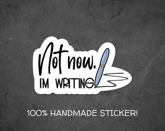 Può includere: Un adesivo bianco con testo nero che dice "Non ora. Sto scrivendo." e una penna blu con una punta gialla che disegna una linea. L'adesivo è su uno sfondo grigio e dice "100% STICKER FATTO A MANO!" in basso.