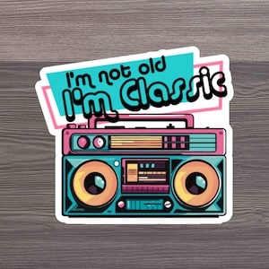 Puede incluir: Un boombox de estilo vintage con el texto "I'm not old I'm Classic" en un esquema de colores rosa, azul y blanco. El boombox tiene una grabadora de casetes rosa y un exterior azul y verde.