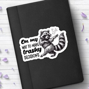 以下が含まれることがあります： いたずらっぽい表情をしたアライグマが描かれた白黒のステッカー。アライグマは後ろ足で立ち、手に星を持っています。「On my way to make trashy decisions」というテキストがアライグマの下に黒文字で書かれています。