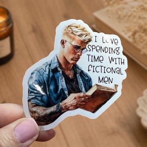 May include: A sticker featuring a man with blonde hair and glasses reading a book. The sticker has the text "I love spending time with fictional men". The man is wearing a blue shirt.
