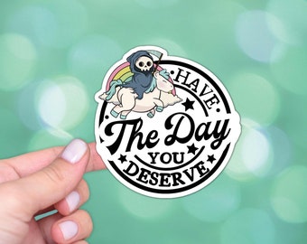 Puede incluir: Una pegatina en blanco y negro con una imagen de dibujos animados de la Muerte montando un unicornio. La pegatina tiene un arcoíris detrás de las figuras y el texto "Have the Day You Deserve." en un círculo negro.