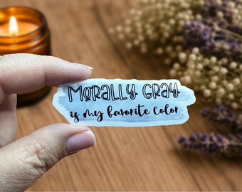 May include: A sticker with the text "Morally Gray is my favorite color" in black script on a light blue background. The sticker is held by a person's hand, with a lit candle and dried flowers in the background.