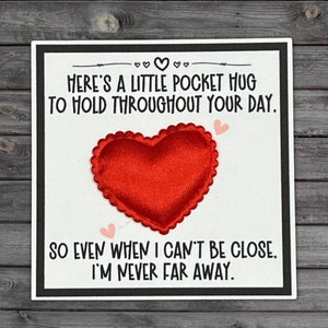 May include: A white card with black text and a red heart-shaped fabric patch. The text reads "Here's a little pocket hug to hold throughout your day. So even when I can't be close, I'm never far away."