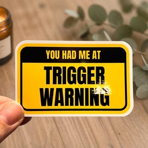 Puede incluir: Una pegatina amarilla y negra con el texto "YOU HAD ME AT TRIGGER WARNING". La pegatina tiene forma rectangular con esquinas redondeadas. Una vela encendida en un tarro de cristal es visible en el fondo.