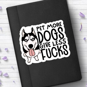 May include: A white sticker with a cartoon husky dog and the text "PET MORE DOGS GIVE LESS FUCKS" in black. The sticker is on a black surface, surrounded by purple flowers and petals.