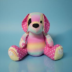 May include: A colourful stuffed dog toy with a rainbow gradient body and pink ears patterned with white hearts. The dog's paws feature a rainbow and cloud design. The toy has a black nose and eyes, against a light blue background.