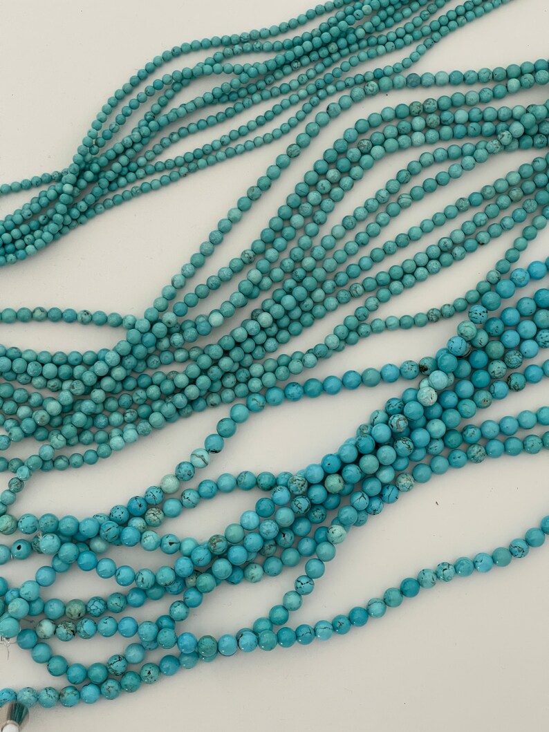 May include: A strand of turquoise beads, with a light blue color, arranged in a loose pile on a white surface.