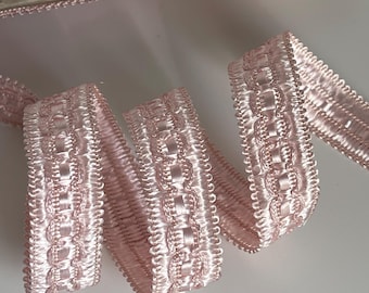 May include: Pale pink decorative trim with a repeating pattern of linked ovals and a scalloped edge. The trim is neatly coiled, with a portion still on a cardboard spool. The material has a slight sheen, suggesting a satin or similar fabric.
