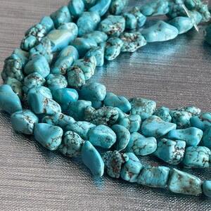 May include: A strand of turquoise gemstone beads, featuring a mix of irregular shapes and sizes. The beads are a light blue color with brown veining and a matte finish.