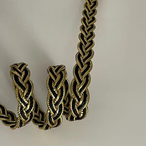 May include: A decorative trim featuring a braided pattern with alternating strands of metallic gold and solid black cord. The flexible trim is coiled in a spiral shape, showcasing the intricate weave and the contrast between the shiny gold and matte black textures throughout the length of the material.