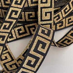 Peut inclure: Ruban noir avec un motif de clé grecque doré à paillettes. Le ruban est enroulé, avec une section déroulée pour montrer le motif. Le motif doré a une texture pailletée, et le ruban est sur une surface blanche.