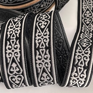 Puede incluir: Cinta decorativa en blanco y negro con un patrón ornamental repetitivo. La cinta está enrollada, mostrando el diseño detallado y la textura. El patrón presenta elementos florales y geométricos estilizados, creando un aspecto clásico y elegante. Ideal para manualidades y adornos.