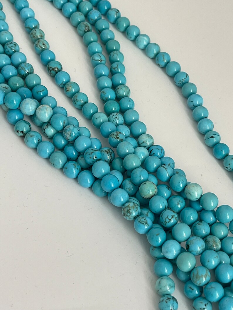 May include: A strand of turquoise beads, featuring a light blue color with dark blue veining. The beads are round and smooth, and they are strung together on a clear thread.