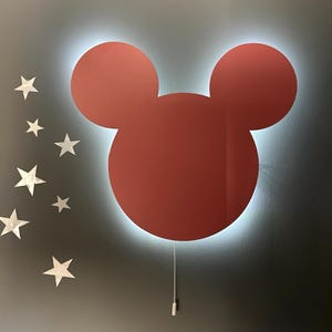 May include: A red Mickey Mouse-shaped night light with a white halo around it. The light is mounted on a wall and has a white cord hanging down.