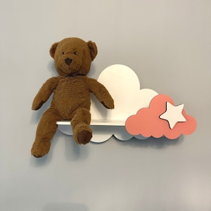 May include: A white and pink cloud-shaped shelf with a brown teddy bear sitting on it. The shelf has a small pink cloud with a white star on it.