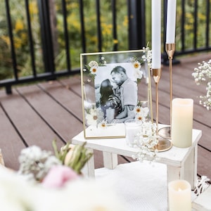May include: A white wooden table with a gold framed picture of a couple kissing. The picture is surrounded by dried flowers. There are two white candles on the table, one in a glass holder and the other on a gold candlestick.