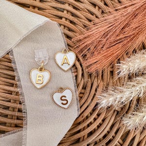 May include: Three white heart-shaped charms with gold trim and gold letters. The letters are A, B, and S. The charms are on a woven wicker surface.