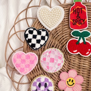 May include: A collection of eight embroidered iron-on patches in various shapes and designs. The patches include a heart with white pearls, a checkered heart, a pink fuzzy heart, a pink disco ball, a purple smiley face with a checkered pattern, a pink flower with a yellow smiley face, a red cherry with a green stem, and a red hot sauce bottle with the text "Hot & Spicy".